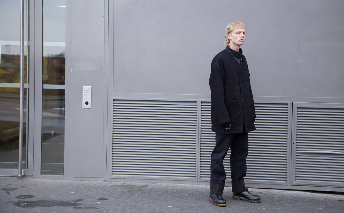 A model in an oversized black jacket and black denim standing before a grey shutter