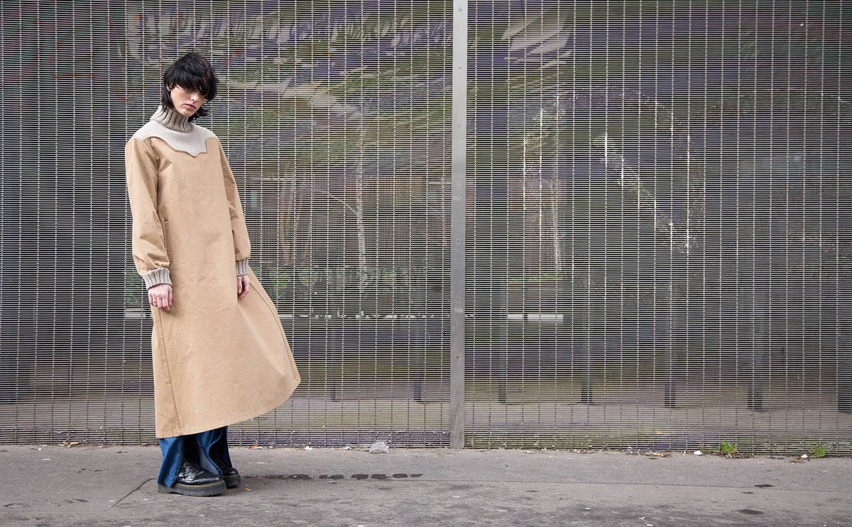 A model in a long beige dress layered over indigo denim against a wire fence