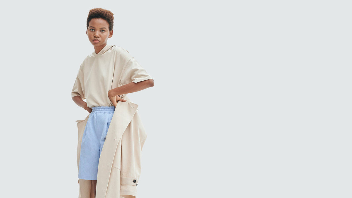 A model in a beige tee and blue shorts with a coat tied at the waist, on white