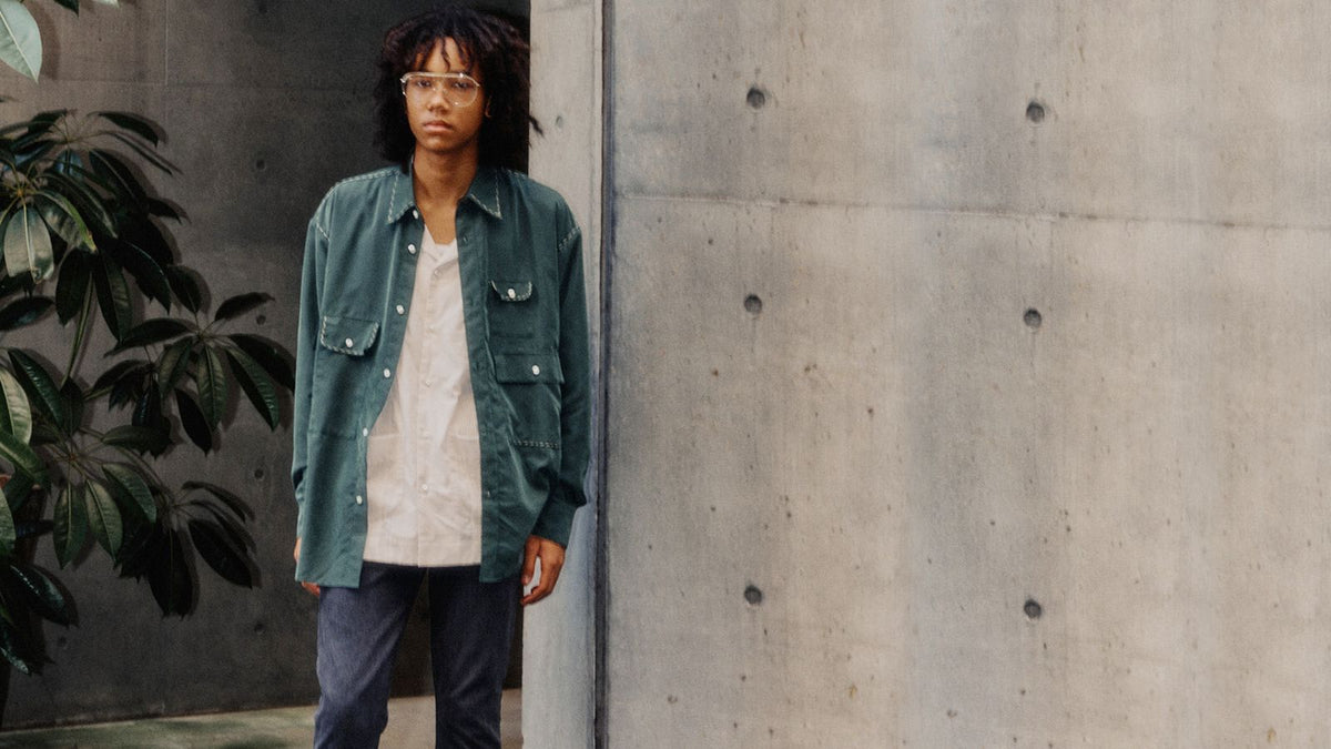 A model in a deep green work shirt and denim beside a concrete wall and foliage