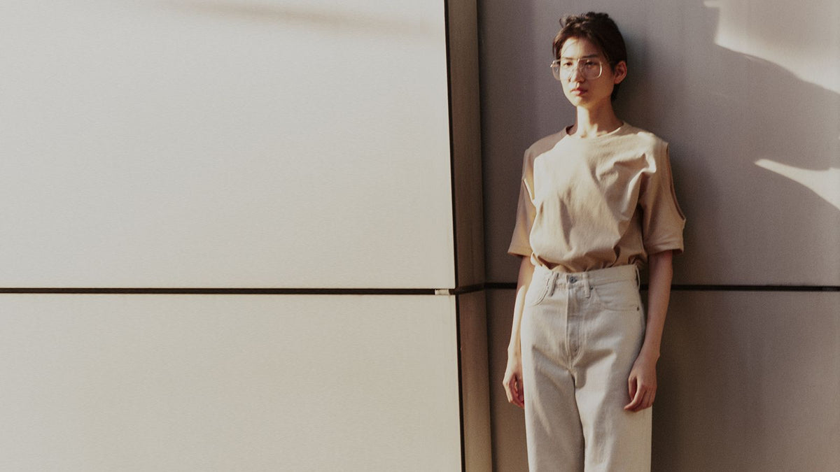 A model in a beige tee and off-white wide denim against a sunlit wall