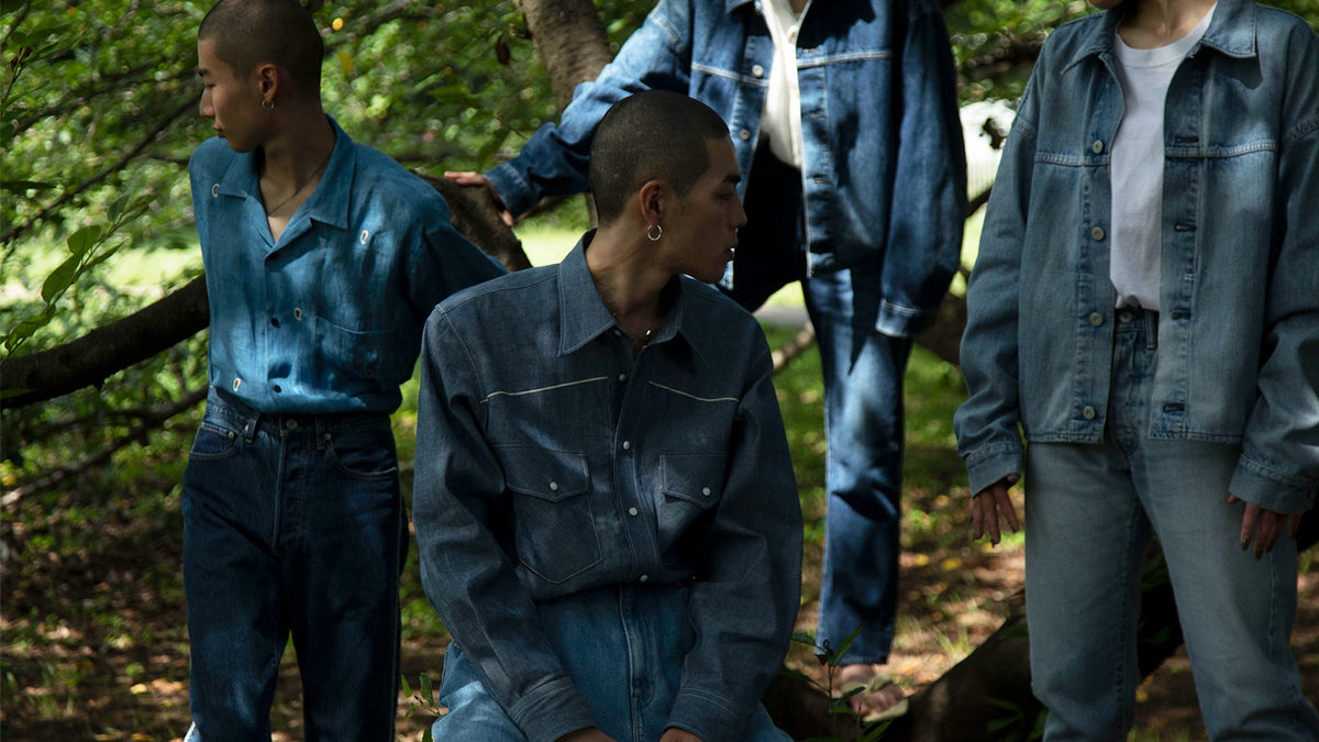 Four models in denim shirts and trousers of varying washes among sunlit trees