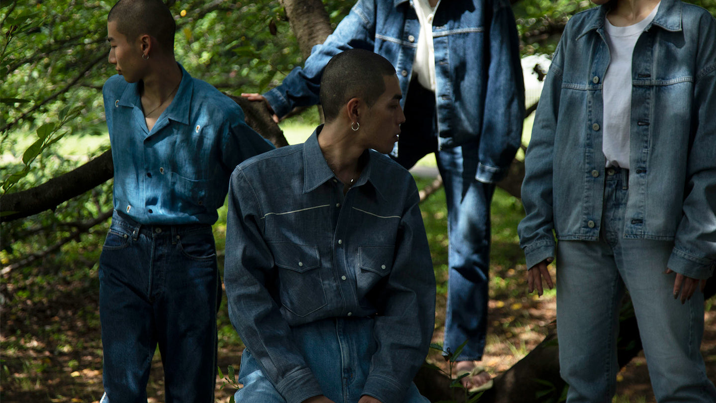 Four models in denim shirts and trousers of varying washes among sunlit trees