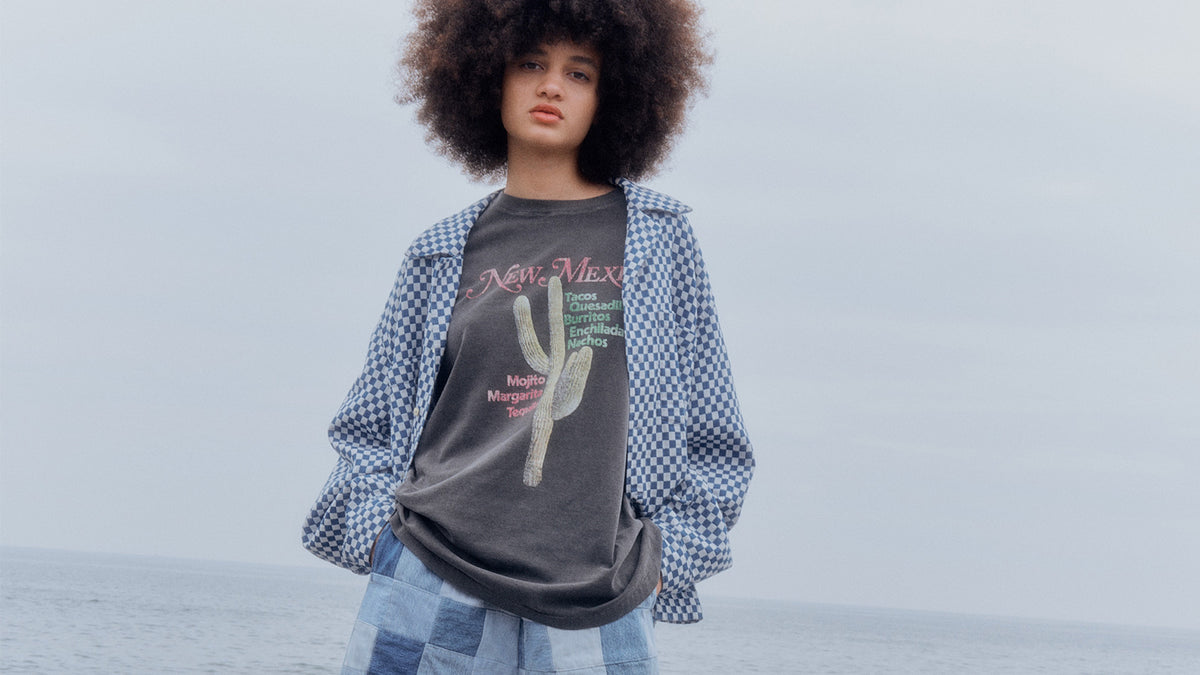 A model in a printed tee under a gingham shirt with patchwork denim, sea behind