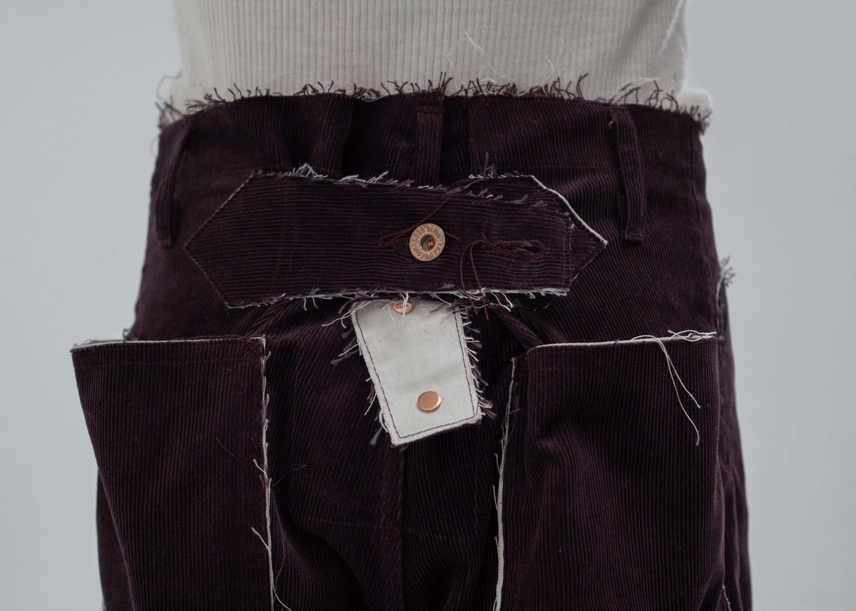 Waist detail of purple corduroy trousers showing a raw-cut waistband, a hexagonal tab, copper buttons and an ecru fly patch.